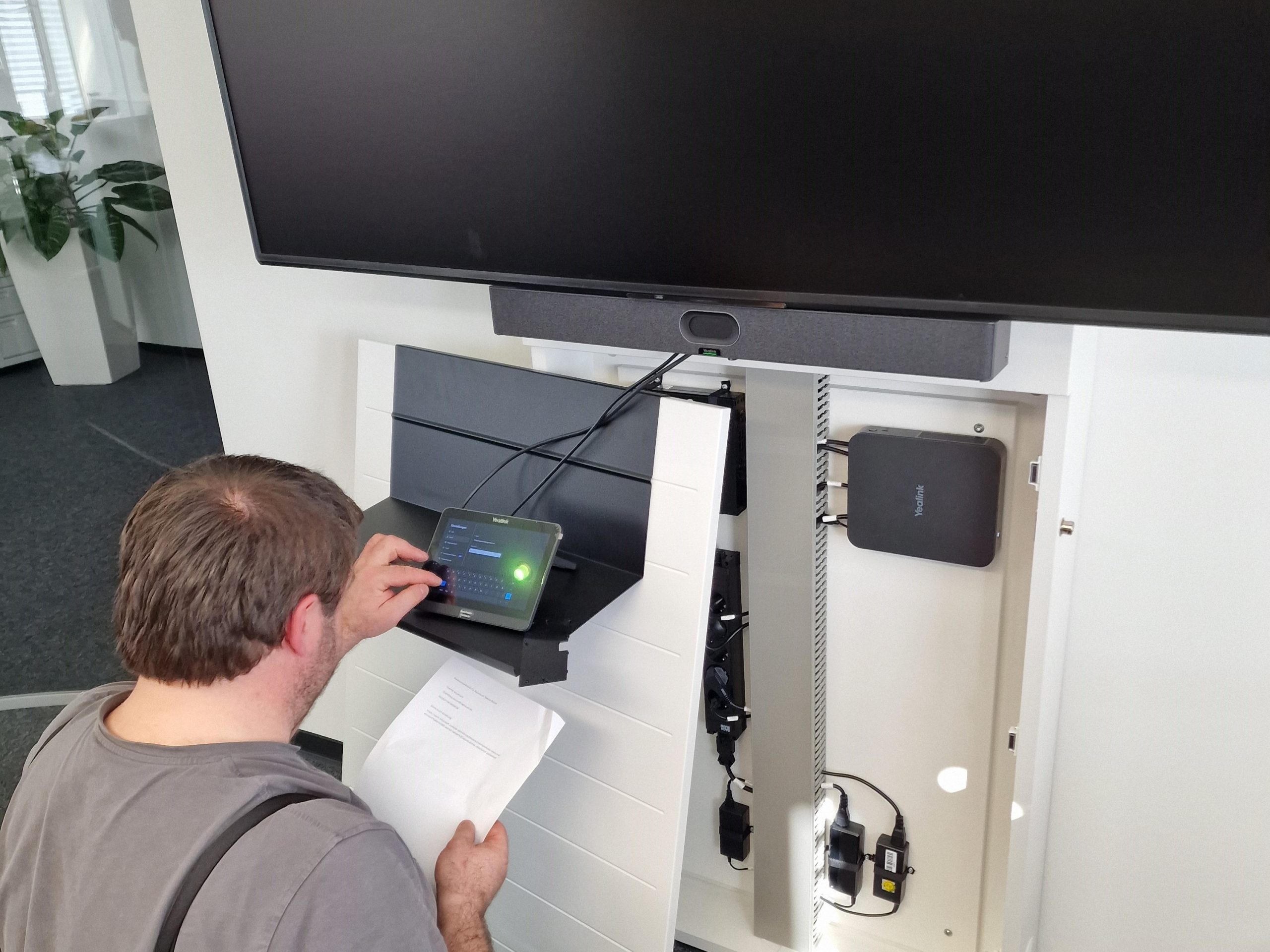 Ein Techniker bedient ein Touchpanel zur Einrichtung eines Konferenzraumsystems, während er ein Blatt mit Installationsdaten in der Hand hält.