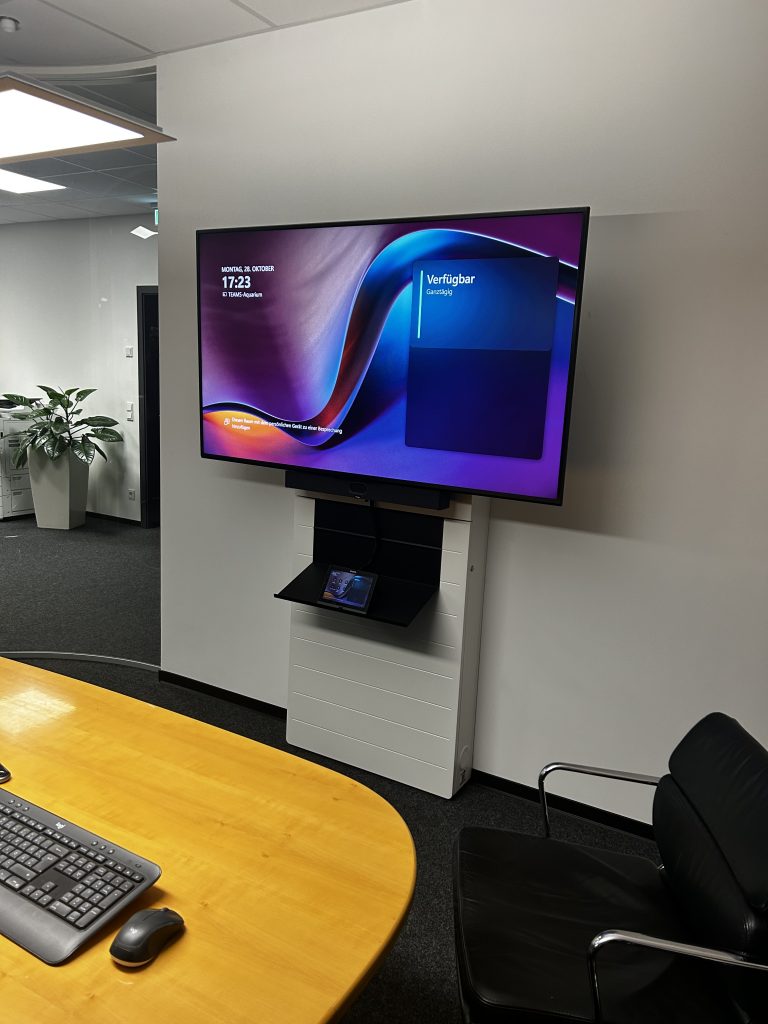 Ein großer Bildschirm mit Videokonferenz-Oberfläche ist über einem integrierten Touchpanel in einem modernen Huddle Room montiert.