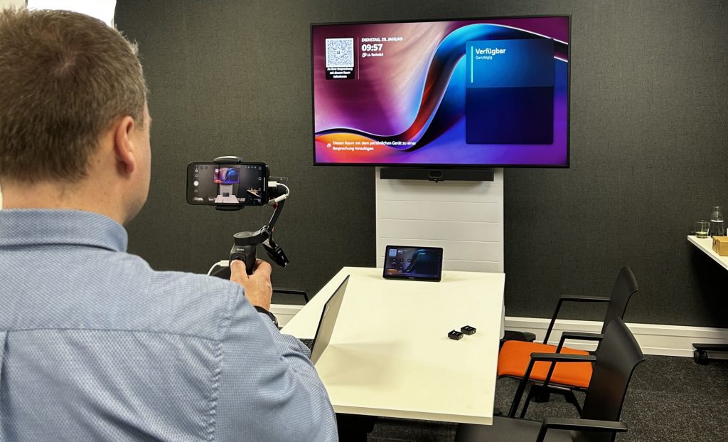 Ein Mitarbeiter filmt mit einem Smartphone-Gimbal einen kleinen Konferenzraum, in dem ein großer Bildschirm mit Meeting-Software, ein Tisch mit Tablet und ClickShare-Buttons sowie ein Wandlautsprecher zu sehen sind.