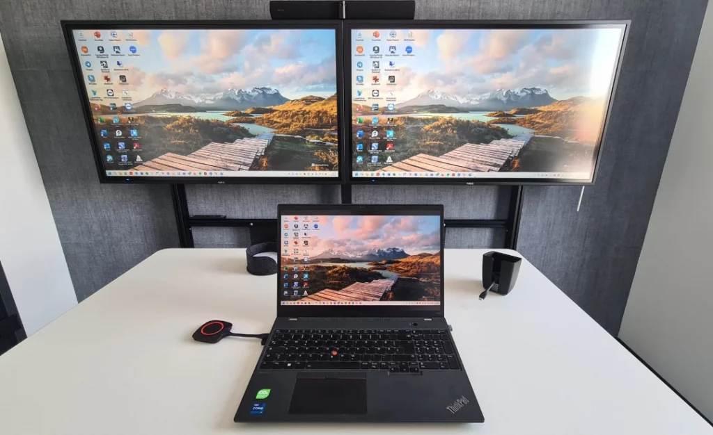 Auf einem höhenverstellbaren Tisch steht im Vordergrund ein aufgeklappter Laptop. Angeschlossen ist ein ClickShare-System, wodurch der Bildschirm automatisch auf die beiden Bildschirme an der Wand gespiegelt wird.