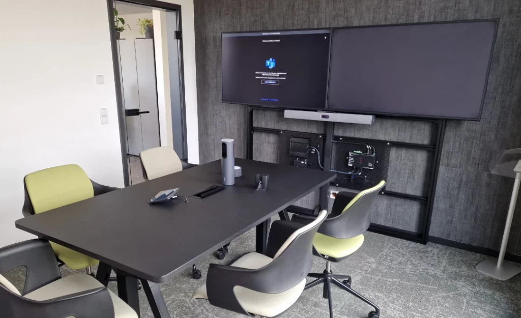 Blick in den Konferenzraum Lech - ein Tisch mit fünf Stühlen, darauf ein Speaker-Tracking-System und ClickShare-System. An der Wand sind zwei große Bildschirme.