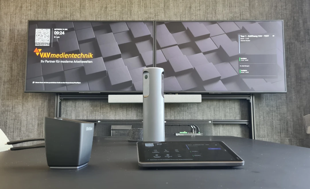 Auf einem Tisch steht ein Speaker-Tracking-System. Im Hintergrund sind andere Wand zwei große Bildschirme angebracht.