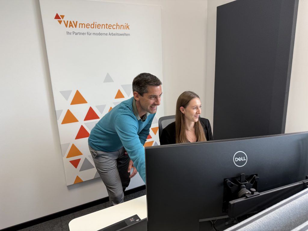 Zwei VAV-Mitarbeitende arbeiten am großen Monitor; der Kollege erklärt etwas, die Kollegin lächelt; VAV-Branding an der Wand.